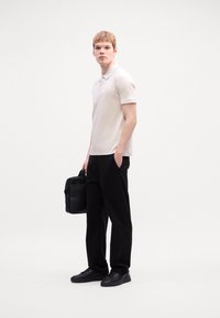 Polo shirt bej deschis cu detalii contrastante, pantaloni negri și adidași negri. Modelul ține o geantă neagră structurată. Design simplu, modern.