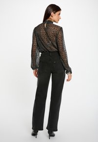 Transparante zwarte blouse met stippenpatroon, gerimpelde kraag, keyhole-detail aan de achterkant, gecombineerd met hoge zwarte jeans en laarzen met hak.