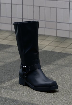 Sort læderstøvle midt på læggen med lav blokhæle og en dekorativ rem med metal spænde rundt om anklen, på en flisebelagt fortov.