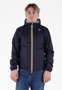 Jeune homme portant une veste à capuche bleu marine avec une fermeture éclair blanche et des poignets élastiques, associée à un jean bleu, debout devant un fond blanc.