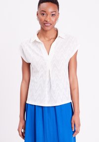 Blouse blanche à manches courtes avec un motif texturé en vagues, design col et coupe décontractée, associée à une jupe bleue.