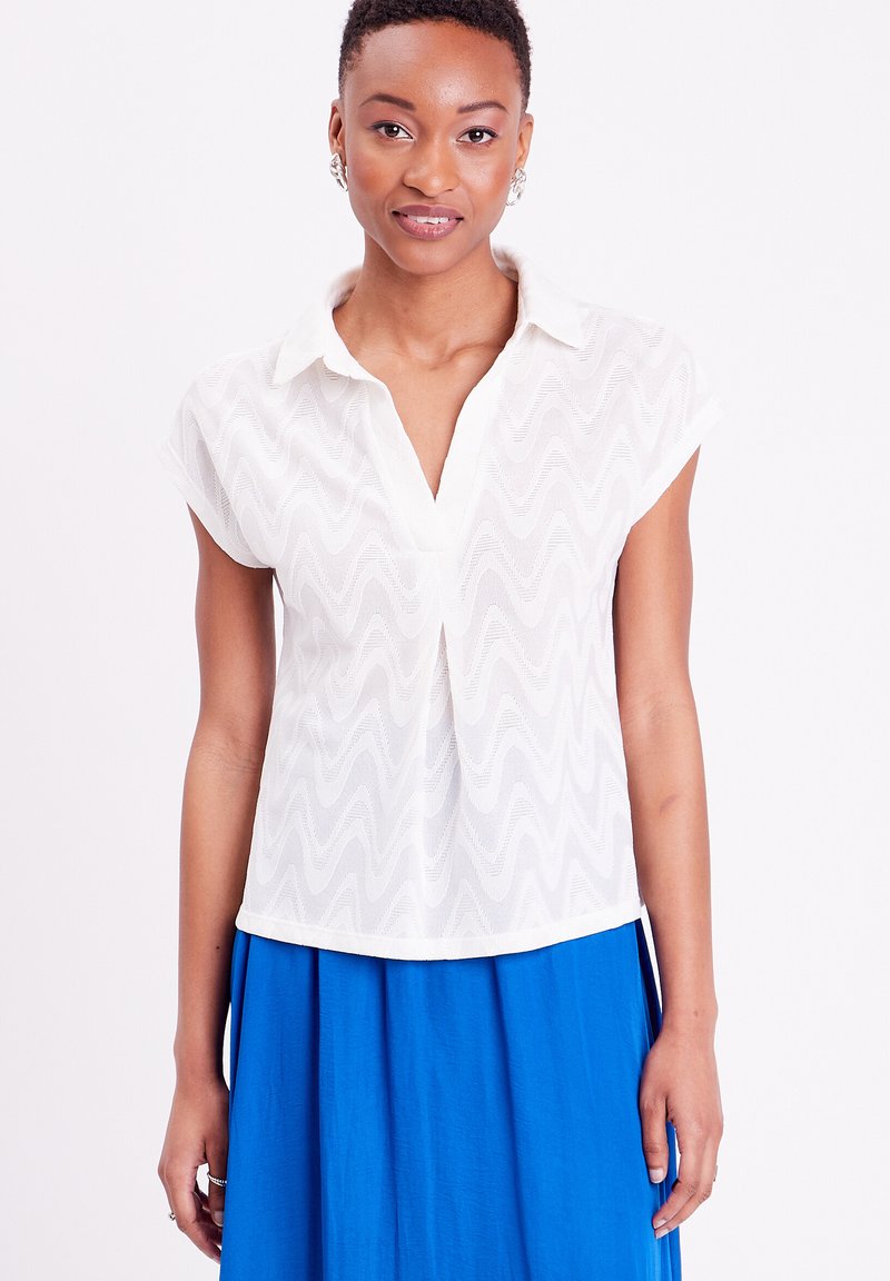 Blouse blanche à manches courtes avec un motif texturé en vagues, design col et coupe décontractée, associée à une jupe bleue.