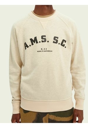 Beigefarbener Pullover mit schwarzem Text "A.M.S. S.C." und kleinerem Text "K. 22 BORN IN AMSTERDAM", getragen von einer stehenden Person.