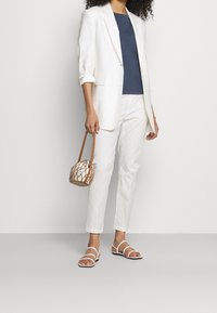 Bílý blazer přes tmavě modrý top, doplněný bílými kalhotami a bílými sandály na podpatku. Drží malou hnědou tašku s pletencovým designem.