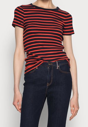 Vrouw die een zwart-rood horizontaal gestreept T-shirt met korte mouwen draagt, lichtjes ingestopt in donkerblauwe spijkerbroek met contrasterende stiksels.
