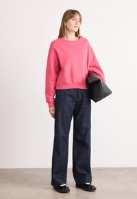 Pink sweatshirt with ribbed cuffs, paired with dark blue wide-leg jeans and black flat shoes. Holding a black bag, set against a light background.