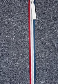 Šedá mikina na zip se třemi kontrastními zipy v červené, bílé a modré barvě, s texturovaným povrchem.