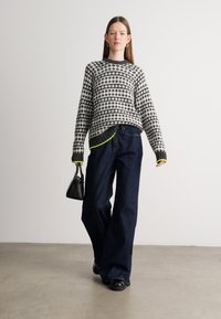 Pulover cu un model gri și negru, cu guler rotund, purtat cu blugi mai largi din denim închis și o geantă mică neagră. Detalii accent în verde neon.