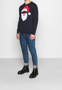 Marinblå tröja med en julgran grafik av jultomten iförd en röd hatt och solglasögon, kombinerad med blå denimjeans och svarta ankelboots.