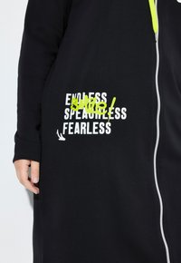 Black garment with white zipper and white text "ENDLESS SPEACHLESS FEARLESS" partly obscured by neon yellow spray paint scribble on sleeve.