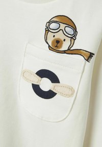 Camiseta de algodón crema con un bolsillo. Presenta un oso de dibujos animados impreso con gafas y un diseño de hélice con suaves acentos beige.