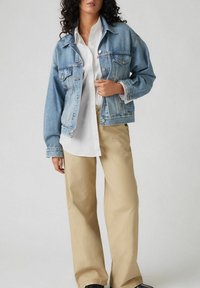 Lichtblauwe spijkerjack met een losse pasvorm, voorzien van borstzakken en knoopsluitingen. Gecombineerd met een wit shirt en beige broek.