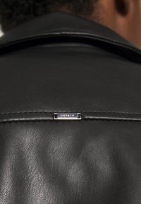 Gros plan sur le dos d'une veste en cuir noire avec une petite étiquette rectangulaire en argent portant l'inscription "MORGAN" près de la couture du col.