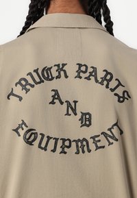 Camicia beige con colletto, caratterizzata da una grafica testuale nera curva che recita "PARTI E ATTREZZATURE PER CAMION". Tessuto morbido.