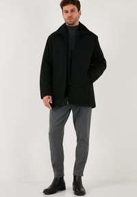 Cappotto nero con zip e colletto, abbinato a pantaloni grigi e stivaletti neri. Il cappotto presenta una texture liscia e opaca.