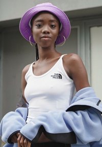 Bílá bezrukávová blůza s černým logem je zkombinována se světle modrou fleecovou bundou. Outfit doplňuje fialová klobouk typu bucket.