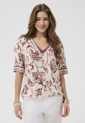 Ung kvinde med langt brunt hår iført en blomsterprintet V-hals bluse med pink og brune mønstre og hvide bukser, smilende mod en ensfarvet baggrund.