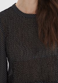 Blusa nera trasparente con pois bianchi, ha un collo rotondo e una leggera texture. Presenta maniche lunghe e un tessuto leggero e fluido.