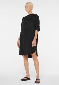 Schwarzes, knielanges Kleid aus leichtem Stoff. Es verfügt über einen runden Ausschnitt, eine durchgehende Knopfleiste, lange hochgerollte Ärmel und seitliche Schlitze.