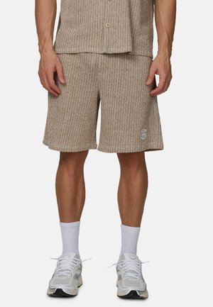 Mężczyzna ubrany w beżowe, prążkowane szorty dzianinowe zestawione z dopasowaną koszulą, białe skarpetki za kostkę i jasnoszare sportowe buty.
