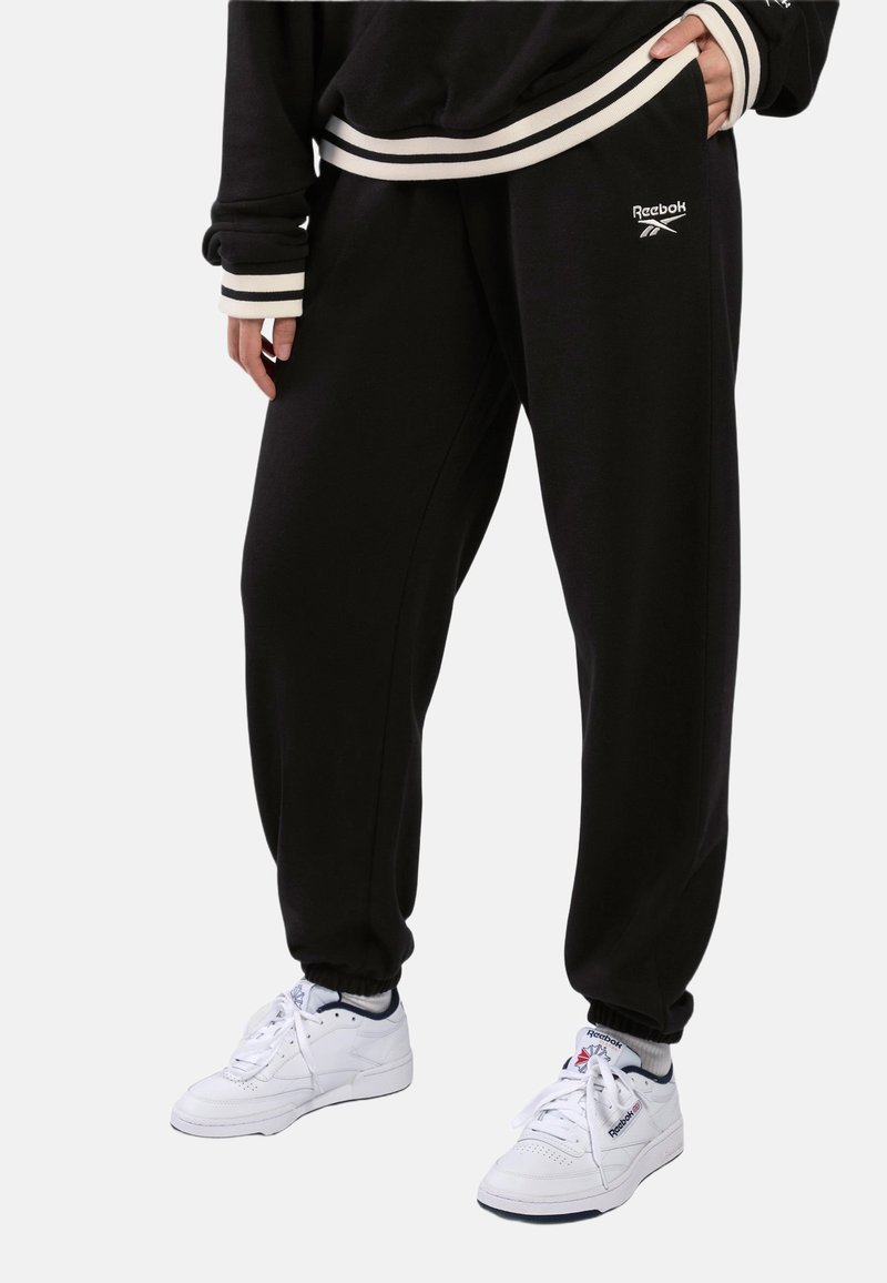 Personne portant un pantalon de survêtement noir Reebok et un sweat-shirt noir avec des poignets rayés blancs, assortis à des baskets blanches Reebok.