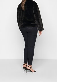 Sort faux fur jakke med lange ærmer, parret med mørkeblå skinny jeans og sorte strophæle, set fra ryggen.