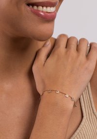 Goldfarbener Armreif mit runden, texturierten Anhängern, die durch eine feine Kette verbunden sind. Besitzt ein zartes Design und eine glatte Oberfläche.