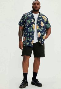 Man som står klädd i en marinblå blommig kortärmad skjorta över en vit t-shirt, svarta shorts, svarta strumpor och svarta snörskor.