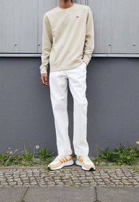 Persona che indossa una felpa beige, pantaloni bianchi e sneaker bianche con strisce arancioni, in piedi contro un muro grigio all'aperto.