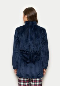 Giacca in velluto a coste blu navy con colletto alto e vita cinchata. Tessuto morbido, maniche lunghe e orlo arrotondato. Nessun hardware visibile.