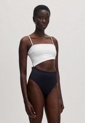 Weißes Cropped-Top mit dünnen Trägern und hochgeschnittenen schwarzen Bikini-Unterteilen; glattes Material, Ausschnitt-Design an der Taille.