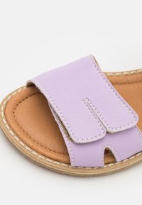 Lawendowe skórzane klapki z szerokim paskiem i wycięciami. Brązowa skórzana wkładka z kontrastowym białym przeszyciem i naturalną podeszwą.