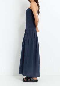 Robe maxi bleu marine sans bretelles avec un corsage froncé et une jupe plissée fluide ; portée avec des sandales noires à double bride.