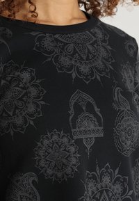 Donna che indossa una maglietta nera con intricati motivi floreali e geometrici di colore grigio scuro, con scollatura tonda e capelli ricci visibili.