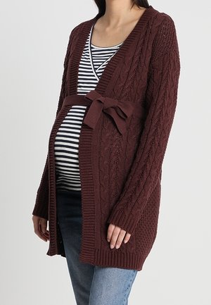 Schwangere Frau trägt einen braunen Strickcardigan mit gebundenem Gürtel über einem schwarz-weiß gestreiften Shirt und Jeans, steht vor einem einfarbigen Hintergrund.