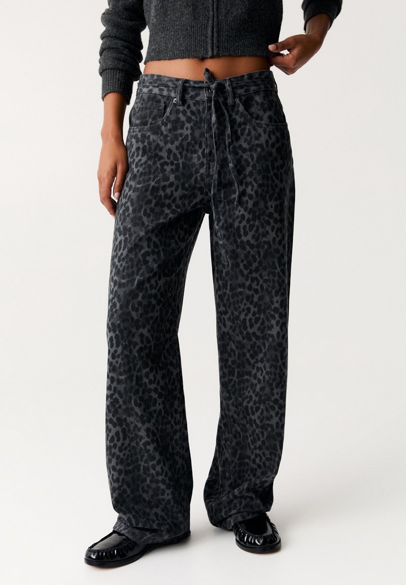 Pantalones de pierna amplia en tela gris oscuro con un patrón de leopardo. Cuentan con una cintura ajustable con cordón y bolsillos laterales. Zapatos negros brillantes.