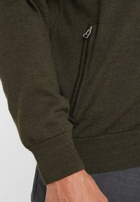 Dunkelgrüner Pullover aus strukturiertem Strick, mit einer seitlichen Reißverschlusstasche und gerippten Bündchen und Saum. Nahaufnahme des Ärmels und des Taschenbereichs.