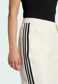 Biele športové nohavice s čiernymi vertikálnymi pruhmi. Majú textúrovaný materiál, elastický pás a bočnú zipsovú vrecko.