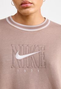Felpa marrone con colletto a costine, logo "NIKE" ricamato e data "1972" in bianco. Texture morbida, vestibilità casual.