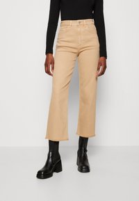 Jeans cortos de pierna ancha color beige con dobladillo deshilachado, que presentan un diseño clásico de cinco bolsillos y cierre de botón, combinados con botas de tacón negras.