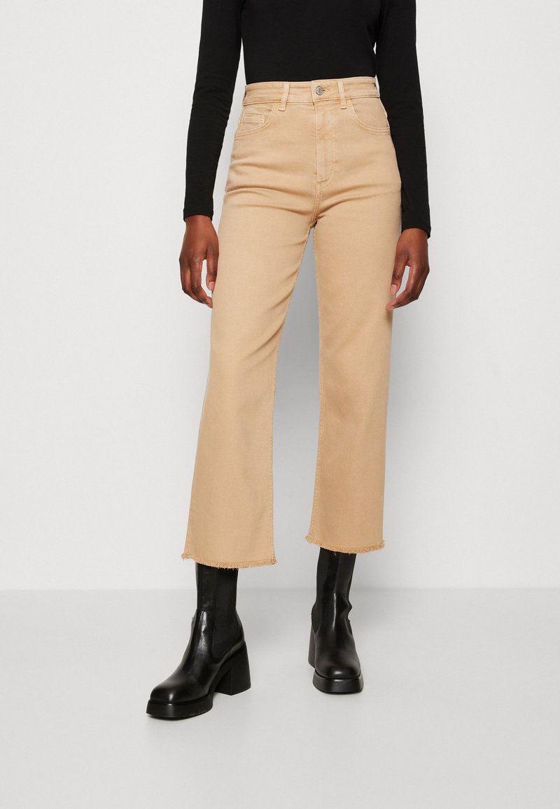 Jeans cortos de pierna ancha color beige con dobladillo deshilachado, que presentan un diseño clásico de cinco bolsillos y cierre de botón, combinados con botas de tacón negras.