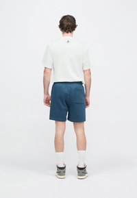 Chemise blanche à manches courtes avec un logo noir dans le dos, associée à un short bleu foncé avec une poche arrière, portée avec des baskets noires et grises.