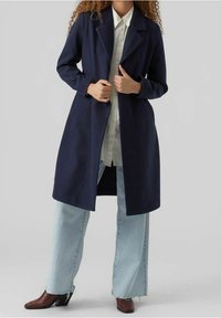 Cappotto di lana blu navy con collo a revers, design fino al ginocchio e tasche laterali, abbinato a jeans a gamba larga di colore chiaro e stivali con tacco marroni.
