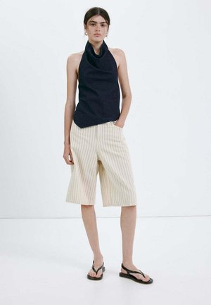 Femme portant un haut à bretelles noir, un short beige à fines rayures jusqu'aux genoux et des sandales noires, debout devant un fond blanc.