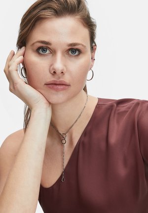 Zilveren ketting met cirkelvormige accenten, gedragen over een bordeauxrode top. Het model heeft halflang haar en steunt haar kin op haar hand.