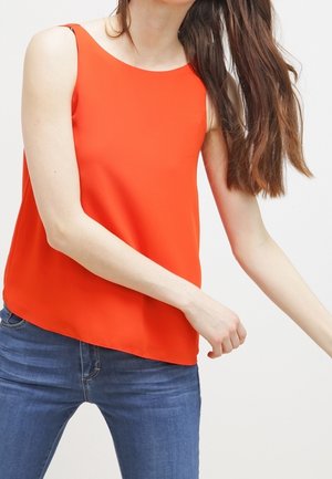 Top rojo sin mangas con cuello redondo, tejido ligero y un dobladillo ligeramente asimétrico, combinado con vaqueros azules.