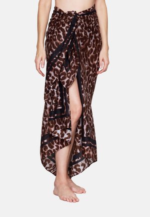 Frau trägt einen braun-schwarzen Sarong mit Leopardenmuster, der an der Taille gebunden ist, zeigt ein Bein, barfuß vor weißem Hintergrund.