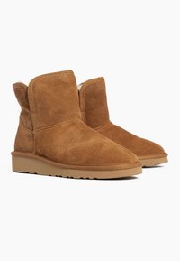 Botas de ante marrón hasta el tobillo con punta redondeada y forro interior suave. Cuentan con una suela de goma plana y detalle de costuras a lo largo de las costuras.