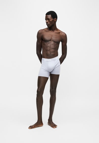 Homme musclé à la peau foncée, debout pieds nus, portant un boxer blanc Lacoste sur un fond clair uni, regardant sur le côté.