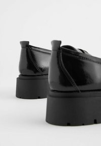 Zwarte schoenen met een glanzende afwerking, voorzien van een chunky zwarte plateauzool en opvallende hiel lussen voor gemakkelijk aan- en uittrekken.